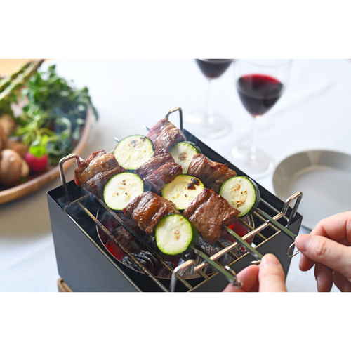 テーブルグリル　TABLETOP　GRILL 黒いテーブルに置かれた金属製のグリルの横からのアングル、奥にはサラダとスパイスのボトルが見える状態。